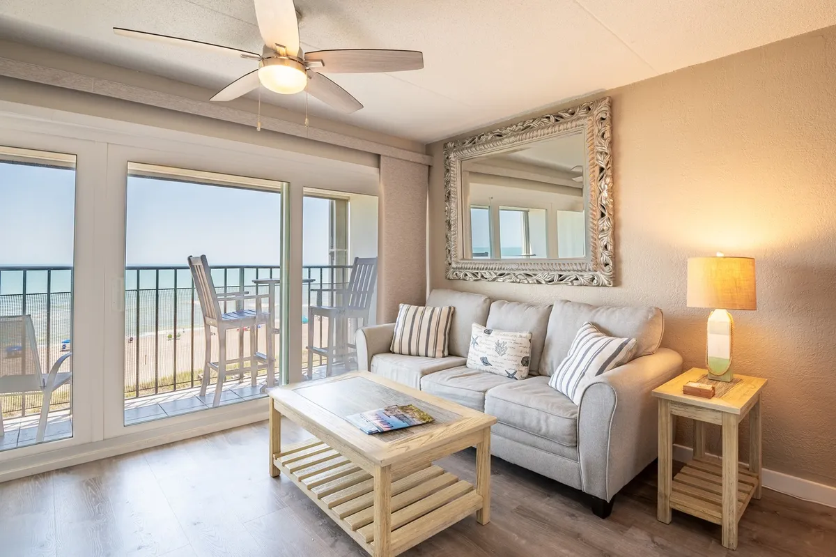 The image showcases a bright, inviting living area with a plush sofa and coffee table, all oriented toward glass doors that open to a private balcony. Through the doors, you can see chairs perfectly positioned for soaking in sweeping ocean views, creating a seamless blend of indoor comfort and seaside serenity.