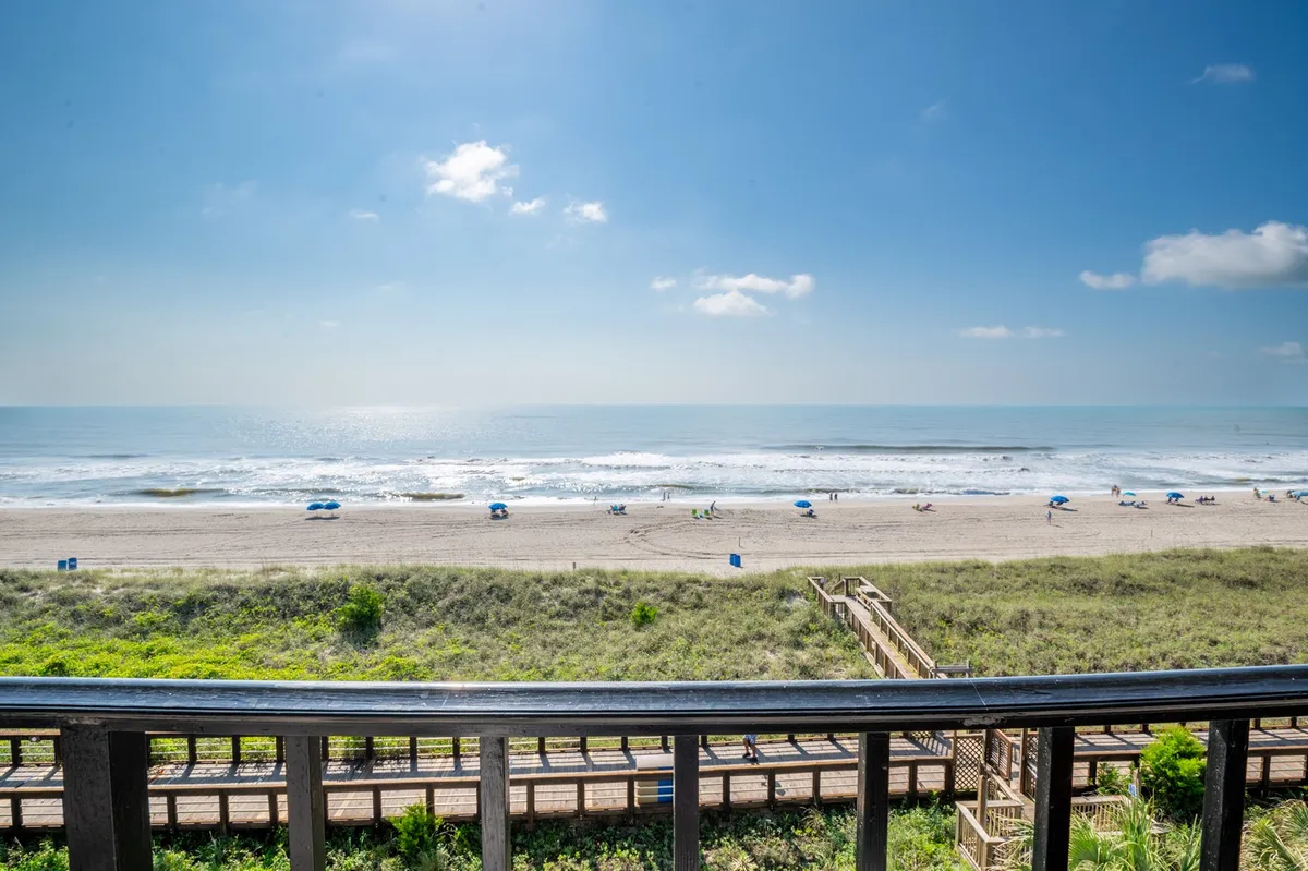 A wide view of the ocean waves rolling onto the sandy shore, with beach chairs lined up along the beach, a boardwalk extending toward the water, and the distant horizon creating a serene backdrop. The image evokes a tranquil, inviting setting perfect for beach strolls and sunset watching.