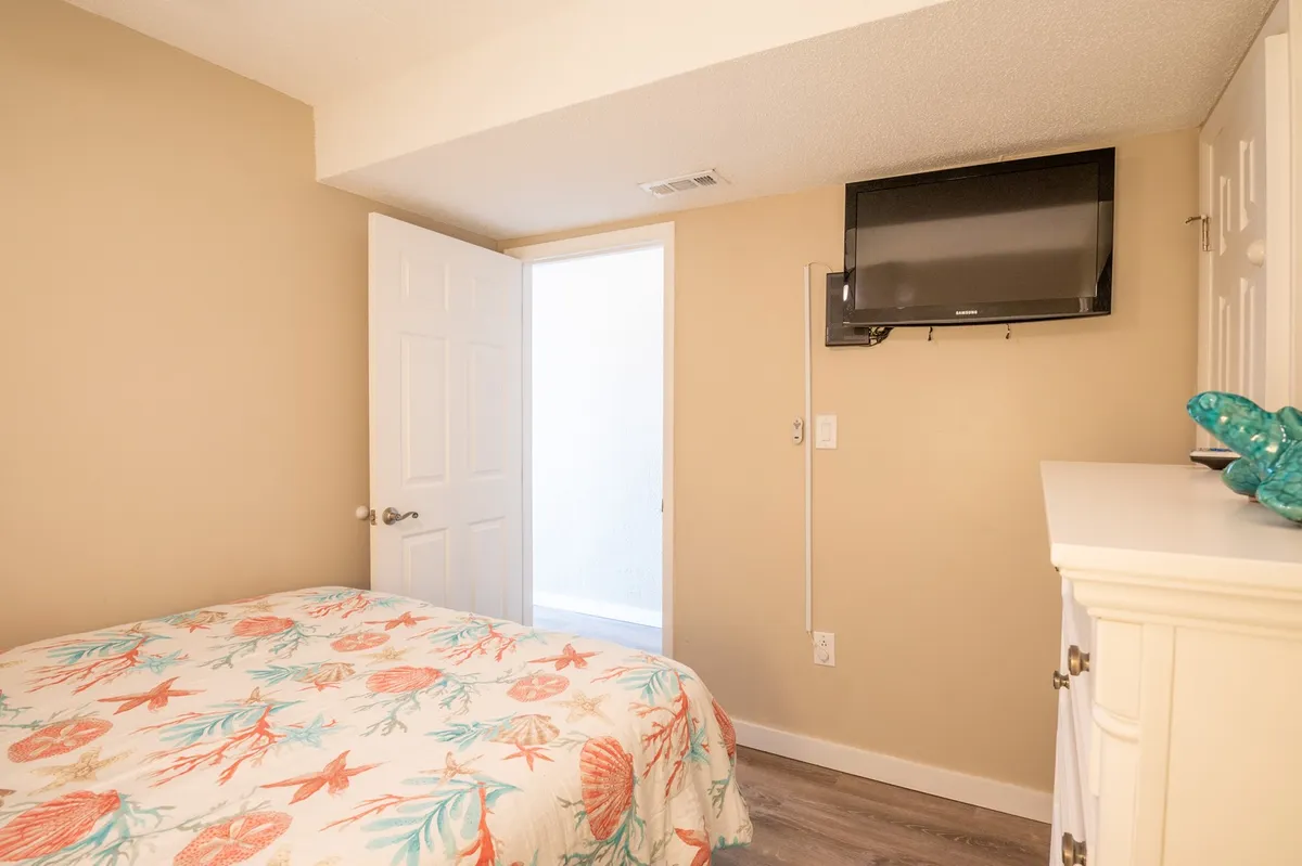 A cozy bedroom with a coral-patterned bedspread, a TV mounted on the wall for private viewing, and a white door leading to an adjoining space. Natural light filters in, making the room feel inviting and comfortable.