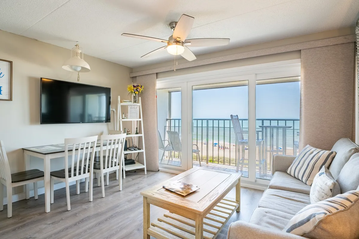 A spacious, sunlit living room with a comfortable sofa positioned to face floor-to-ceiling windows that frame a stunning ocean view. A dining table for four sits conveniently nearby, creating an inviting space for meals or socializing. The overall atmosphere is cozy and peaceful, with décor that enhances the bright, airy feel of the room.