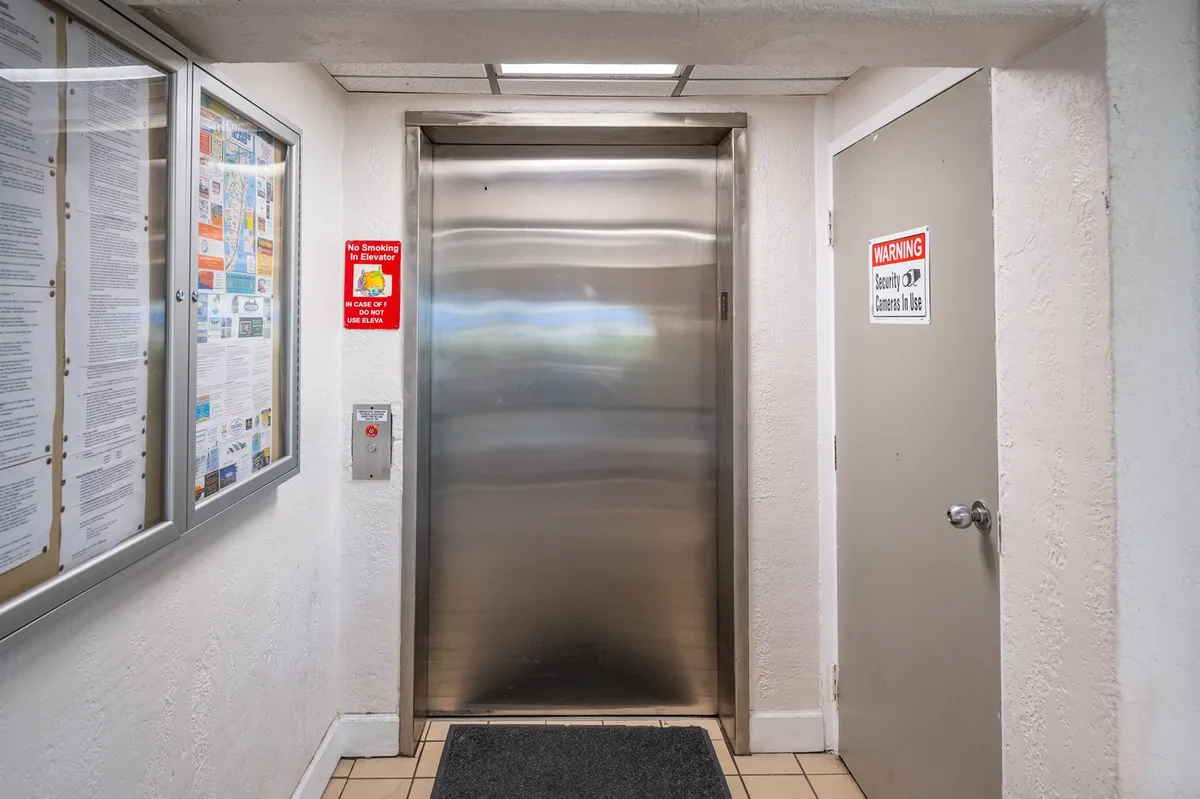 A bright, neutral interior image focusing on the elevator door, call button, and a nearby information board. The scene suggests a point of arrival or departure and subtle moments of community interaction.