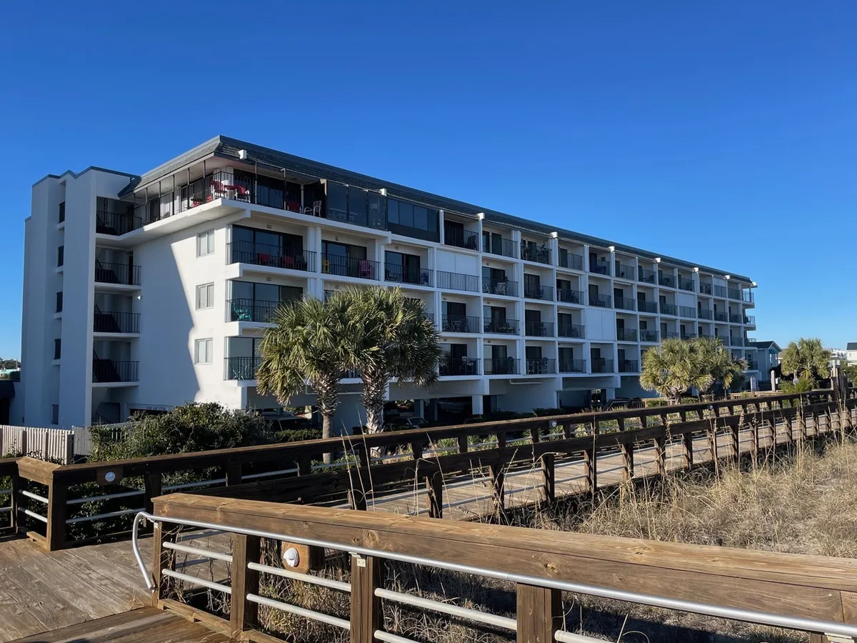 This exterior image showcases the modern architecture of the property with palm trees and a prominent wooden boardwalk leading towards the beach. The scene radiates a welcoming, sunny atmosphere and highlights the direct boardwalk access, echoing the section’s emphasis on proximity to the Carolina Beach Boardwalk and outdoor fun.