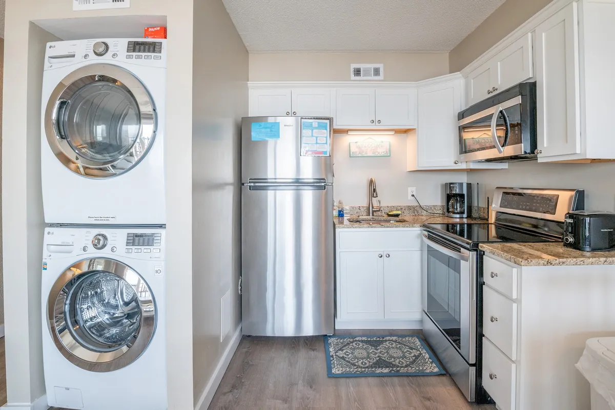 The image displays a practical kitchen area with a washer and dryer, stainless steel refrigerator, granite countertops, stove, and microwave. The appliances are within close reach, highlighting the space's multi-functionality and homey atmosphere.