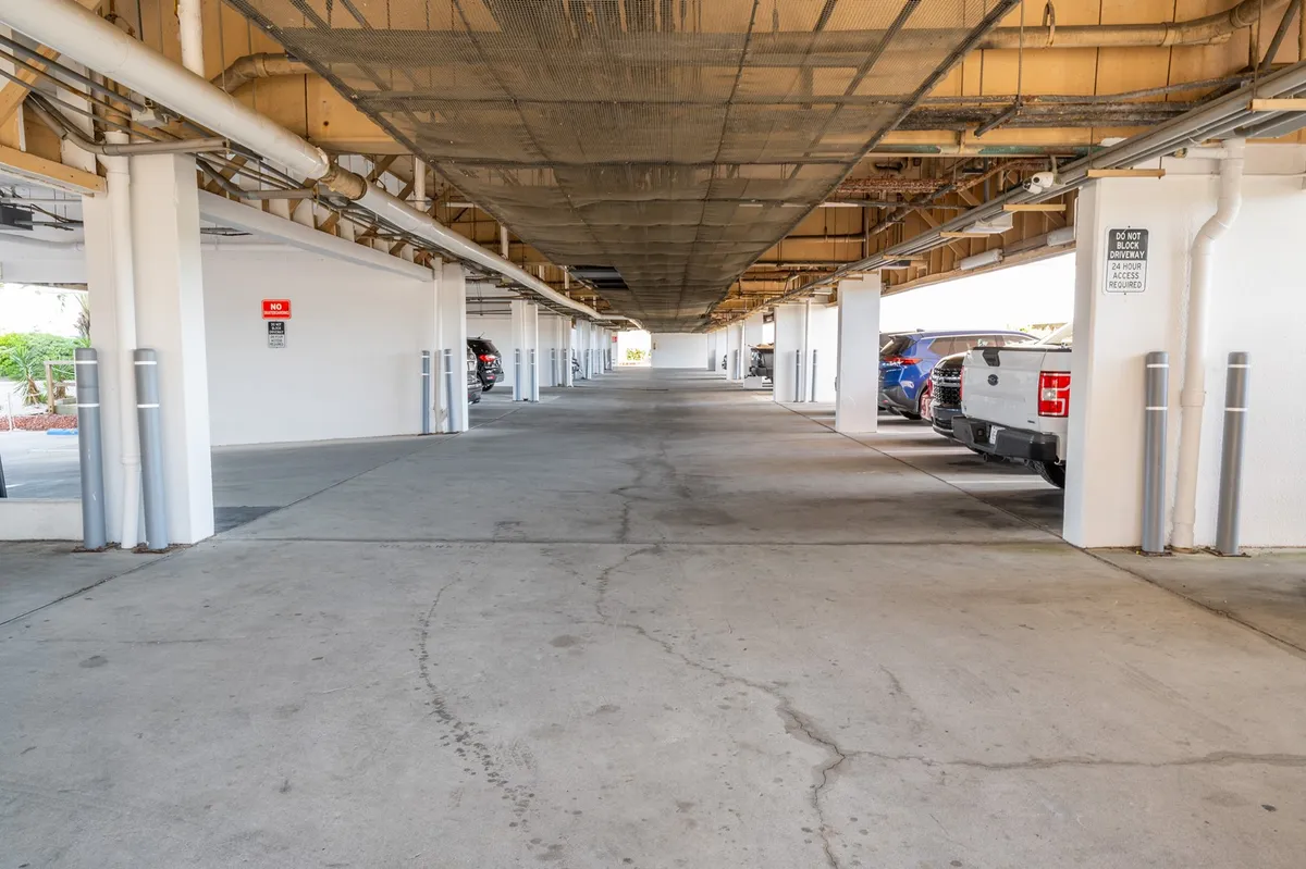 An interior shot of the covered parking area beneath the building, with parking spaces, ceiling structure, and supporting columns visible. The scene feels neutral and functional, highlighting the secure, sheltered aspect of resident parking.