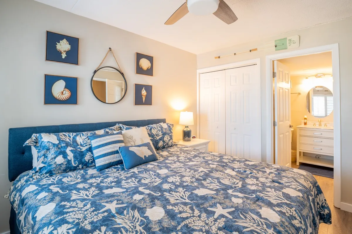 A serene bedroom with a bed dressed in blue patterned bedding, decorative shell artwork above the headboard, a nightstand with a lamp, and a wall mirror. The space is bathed in gentle natural light, creating a calm and inviting atmosphere.