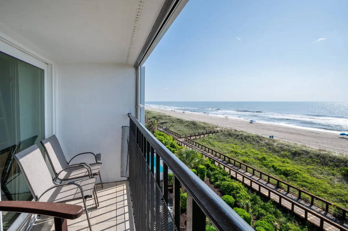 A tranquil balcony scene overlooking the beach and boardwalk, with outdoor chairs positioned for relaxing and taking in the ocean views. The image conveys the possibility of watching fireworks, sunrises, and the bustle of boardwalk life from the comfort of your condo.
