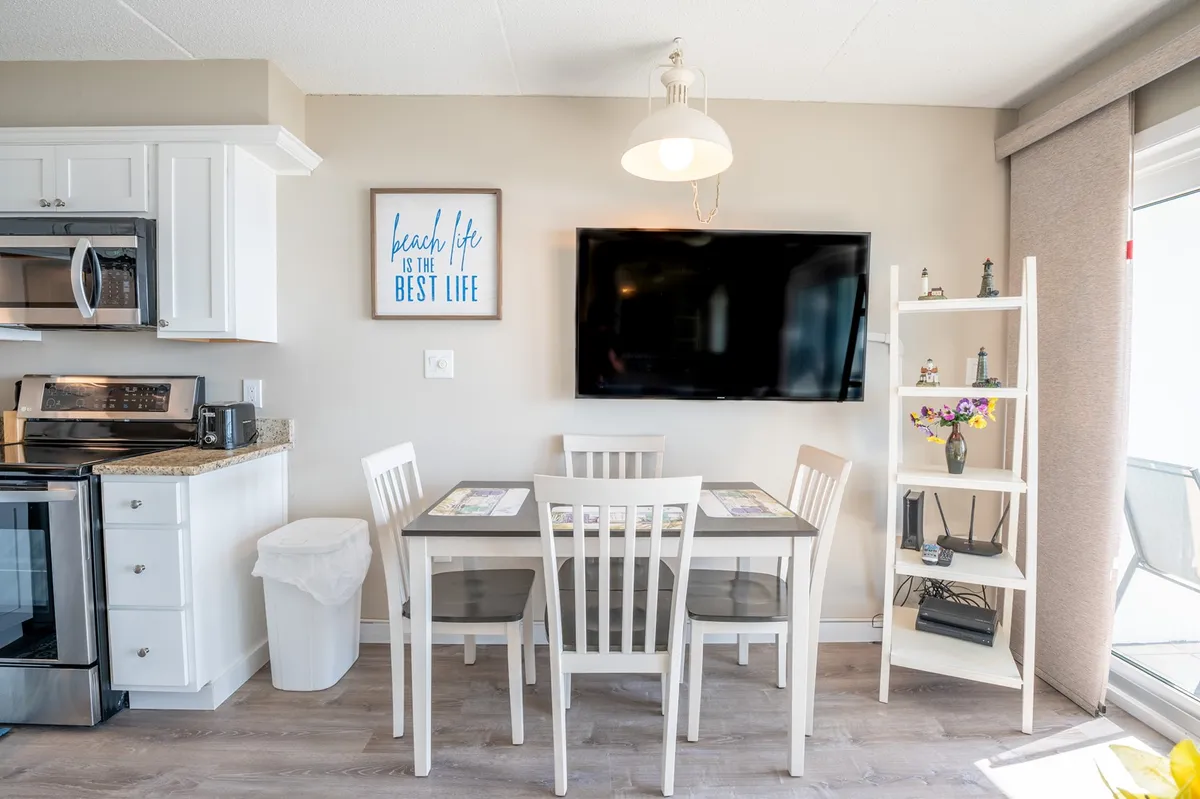 A cheerful dining area adjoining the kitchen, featuring a dining table, TV, kitchen appliances, decorative sign, and bookshelf. The setup invites family meals, game nights, and relaxed gatherings, directly adjacent to the kitchen space.