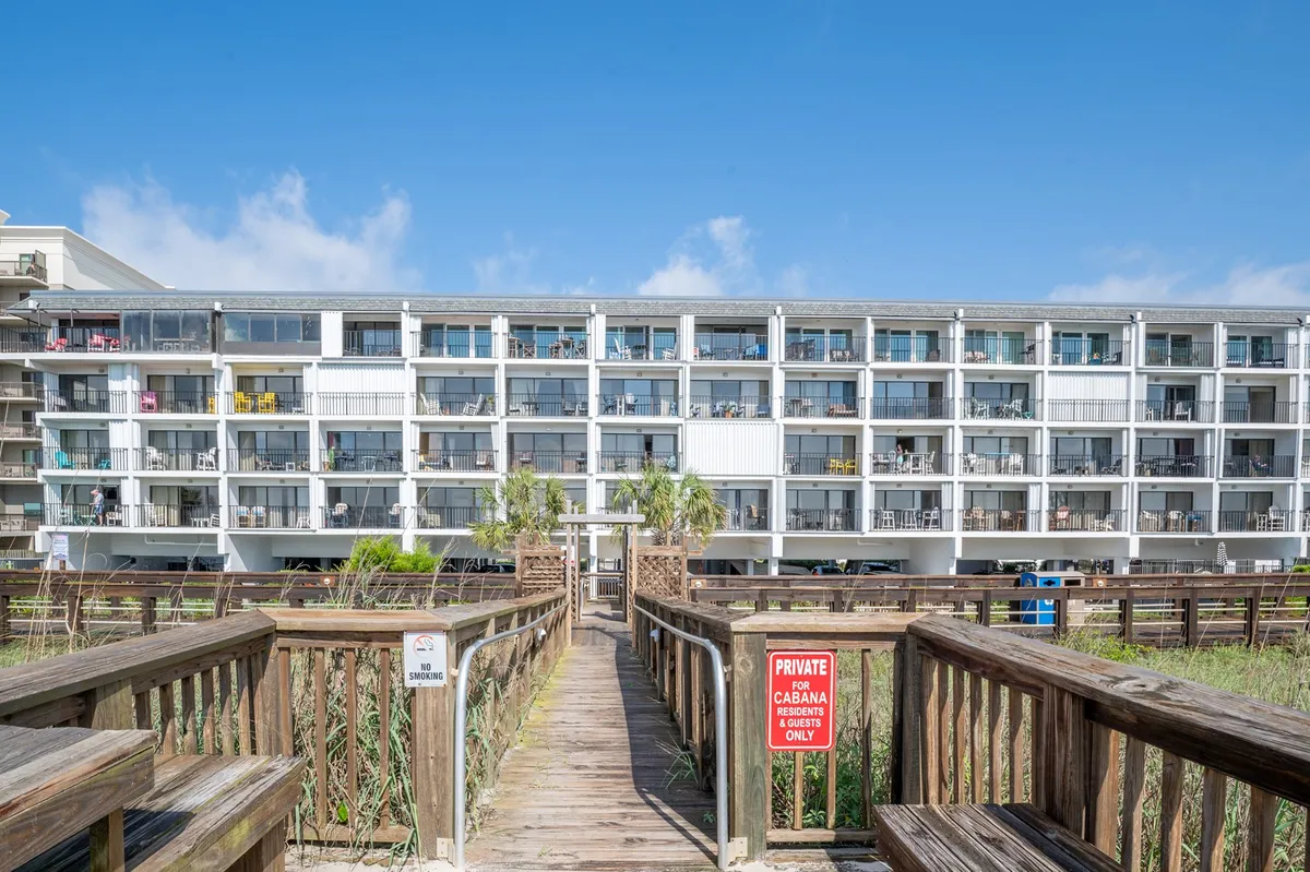 This image shows a wooden boardwalk leading from the building through landscaped grounds with palm trees towards the beach. The multi-story condo building, balconies, and prominent signage reinforce the sense of private, secure access to the shore.