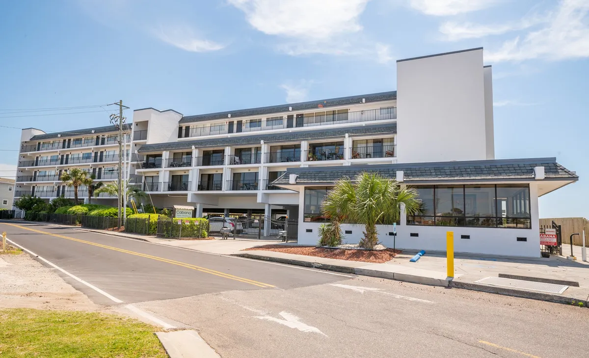 A welcoming exterior of the property featuring the hotel facade with balconies shaded by palm trees, a visible parking area, and hints of sunset light reflecting off the building. The scene is lively yet relaxed, suggesting a social atmosphere ideal for group gatherings or couples.
